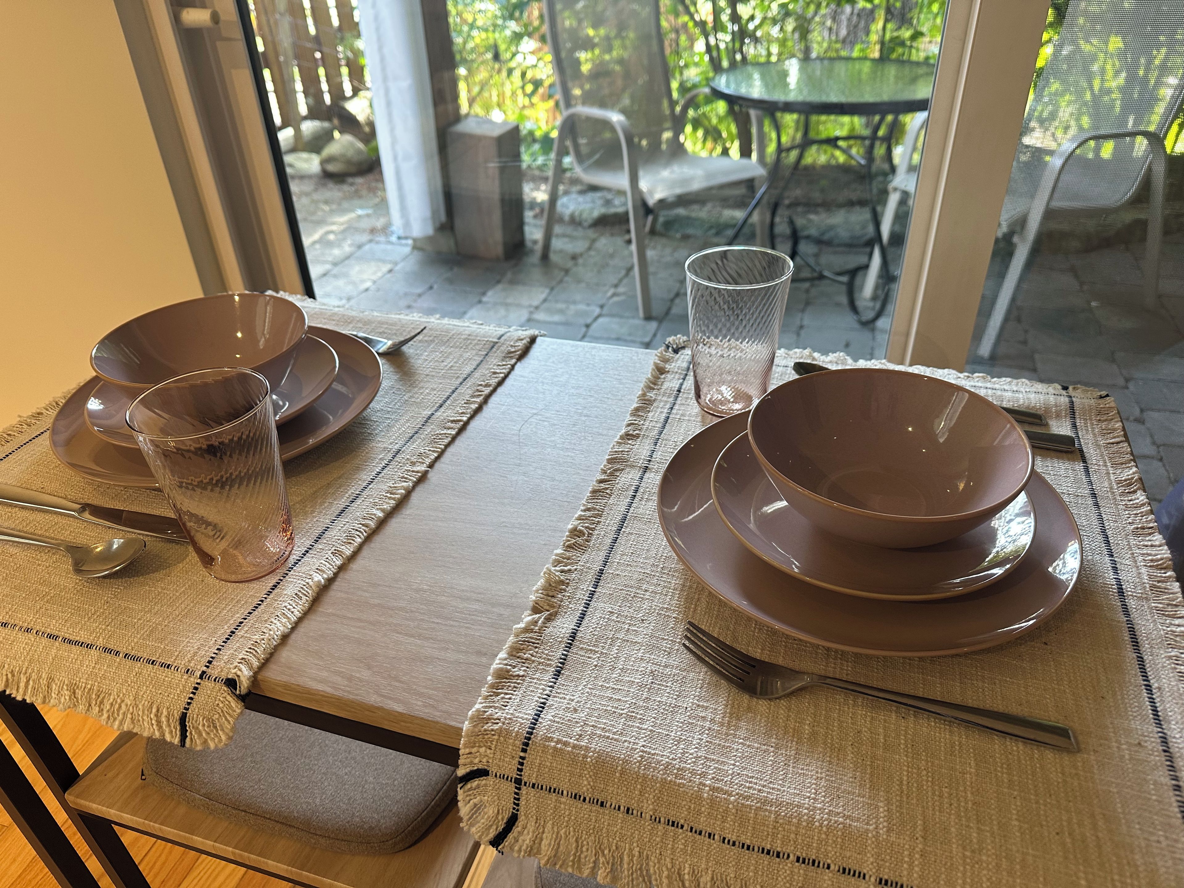 Une table dressée avec des assiettes marron, un verre et des couverts, donnant sur des meubles d’extérieur et de la verdure, le tout visible à travers une porte vitrée.