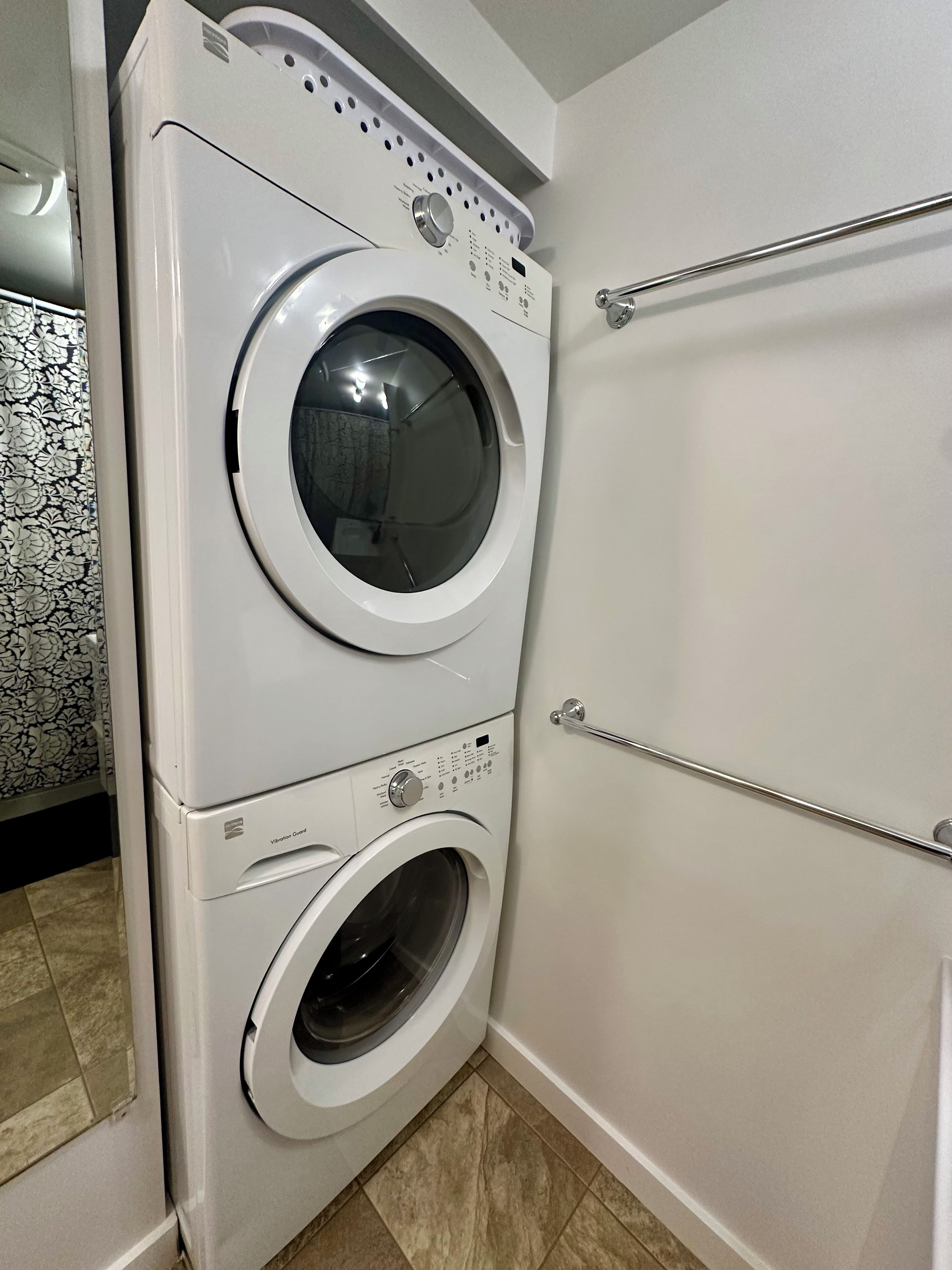 Lavadora y secadora apiladas en un baño con piso de baldosas y un perchero.