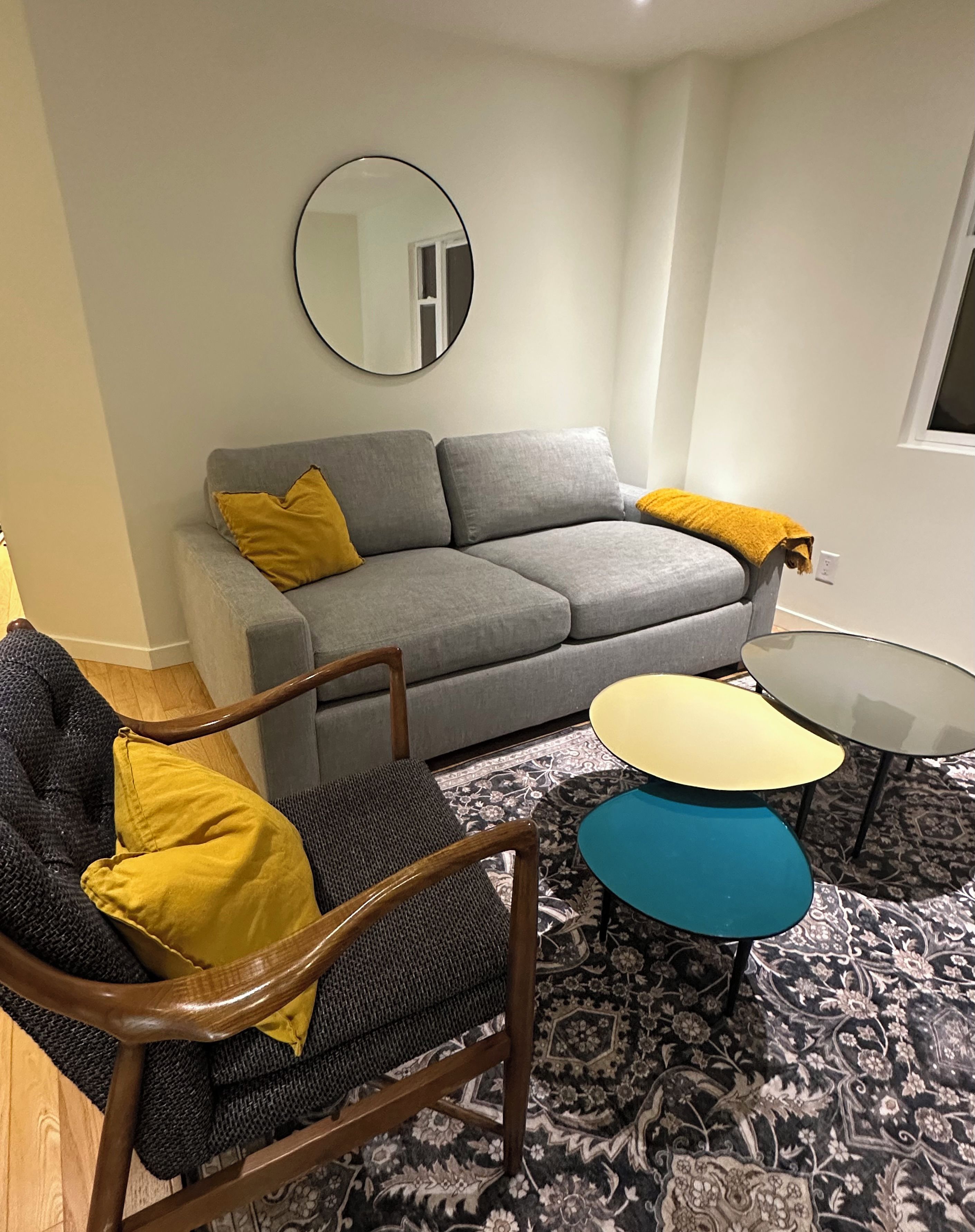 Un salon avec un canap é gris, un miroir rond, une chaise en bois, une petite table et un tapis à motifs.
