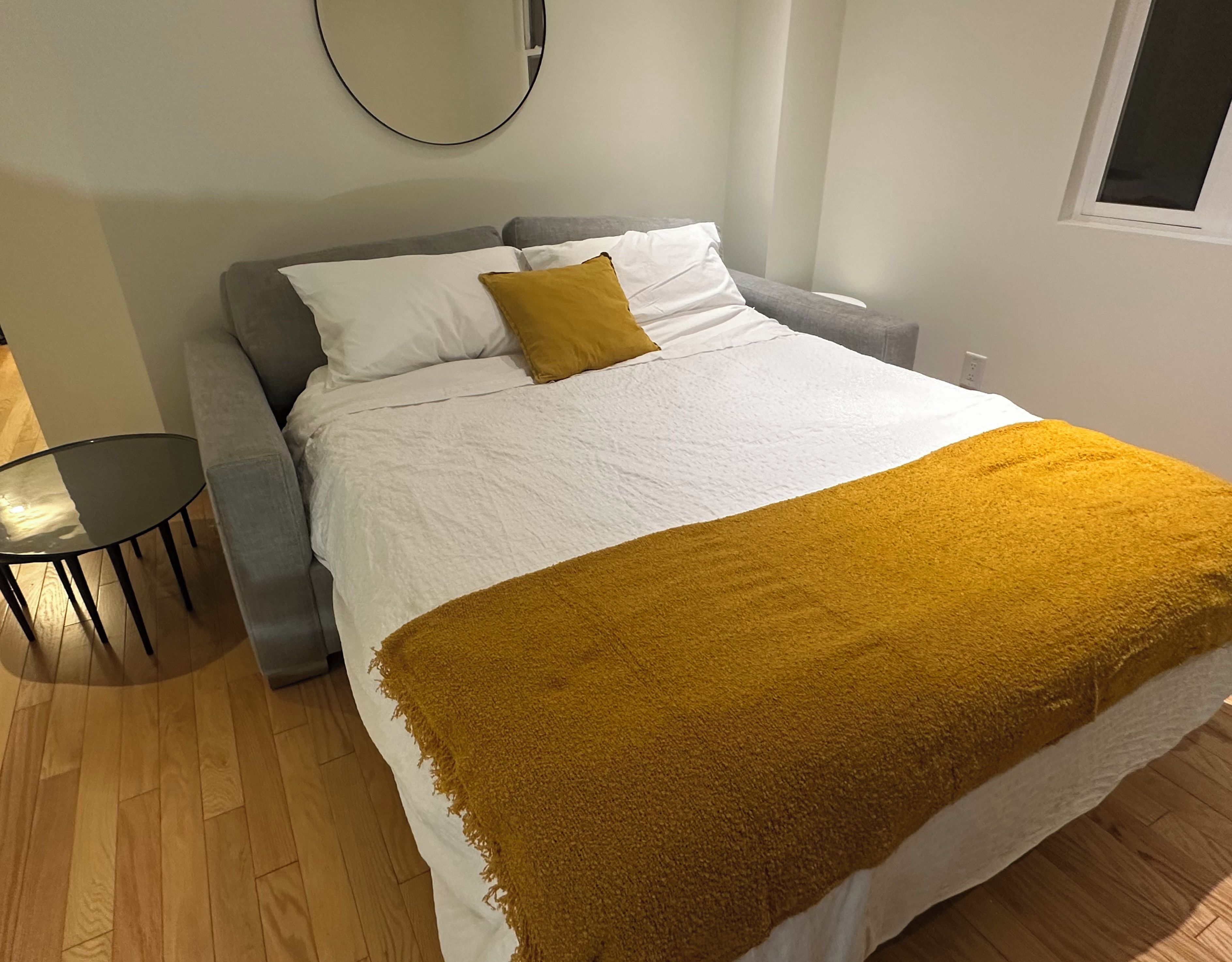 Une chambre à coucher avec un lit, un canapé gris, une petite table ronde et une fenêtre.