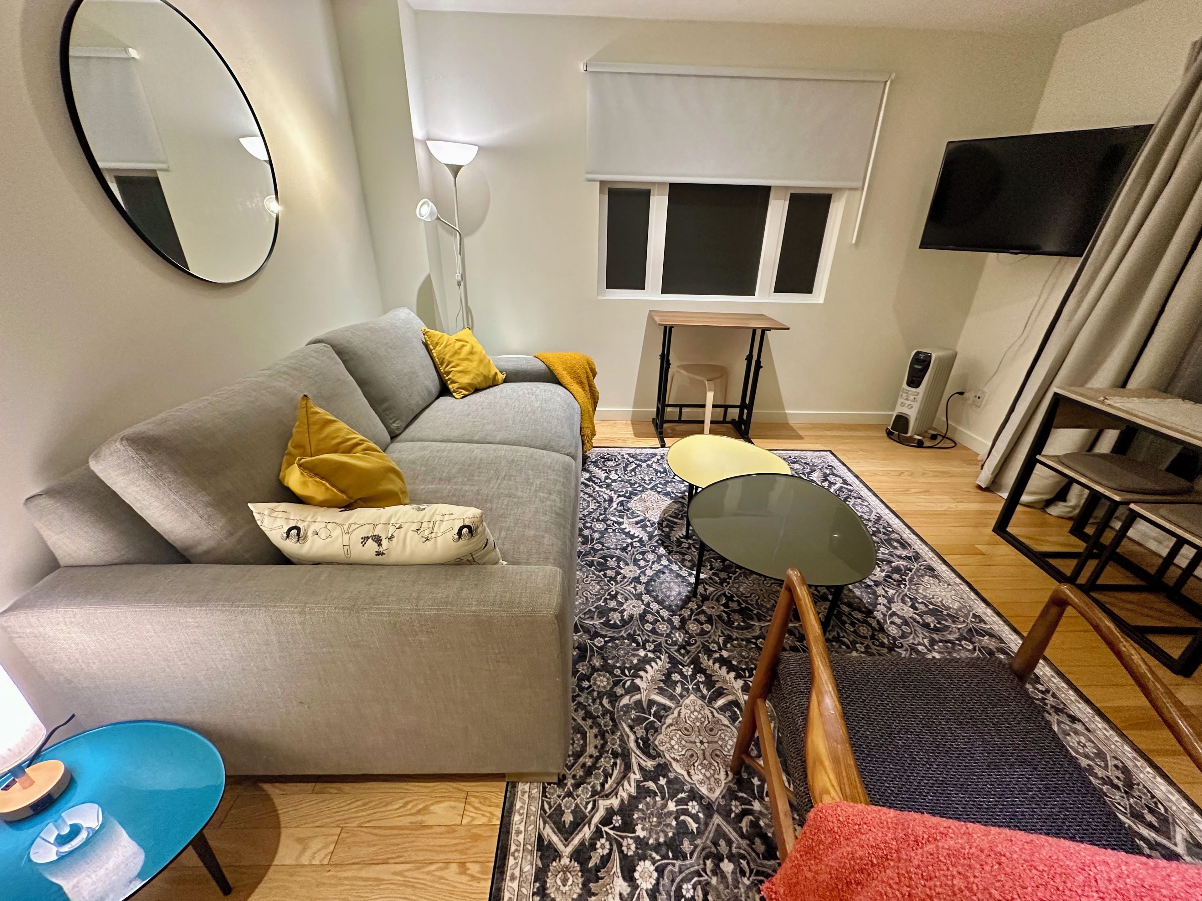 Ein modernes Wohnzimmer mit einem grauen Sofa, einem kleinen runden Tisch, einem gemusterten Teppich und einem Fernseher an der Wand.