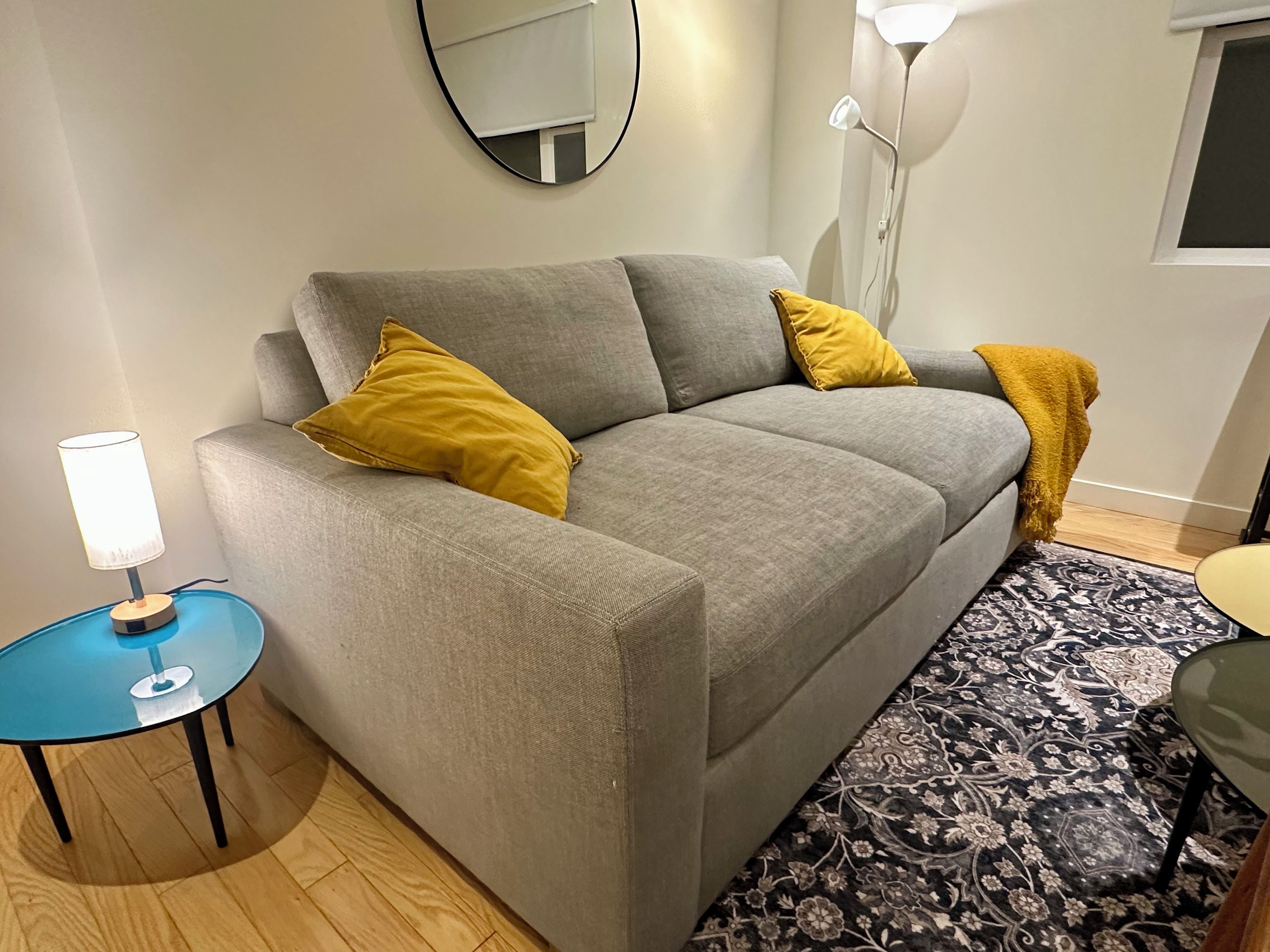 En grå soffa med två gula kuddar, ett litet blått bord med en lampa och en mönstrad matta.