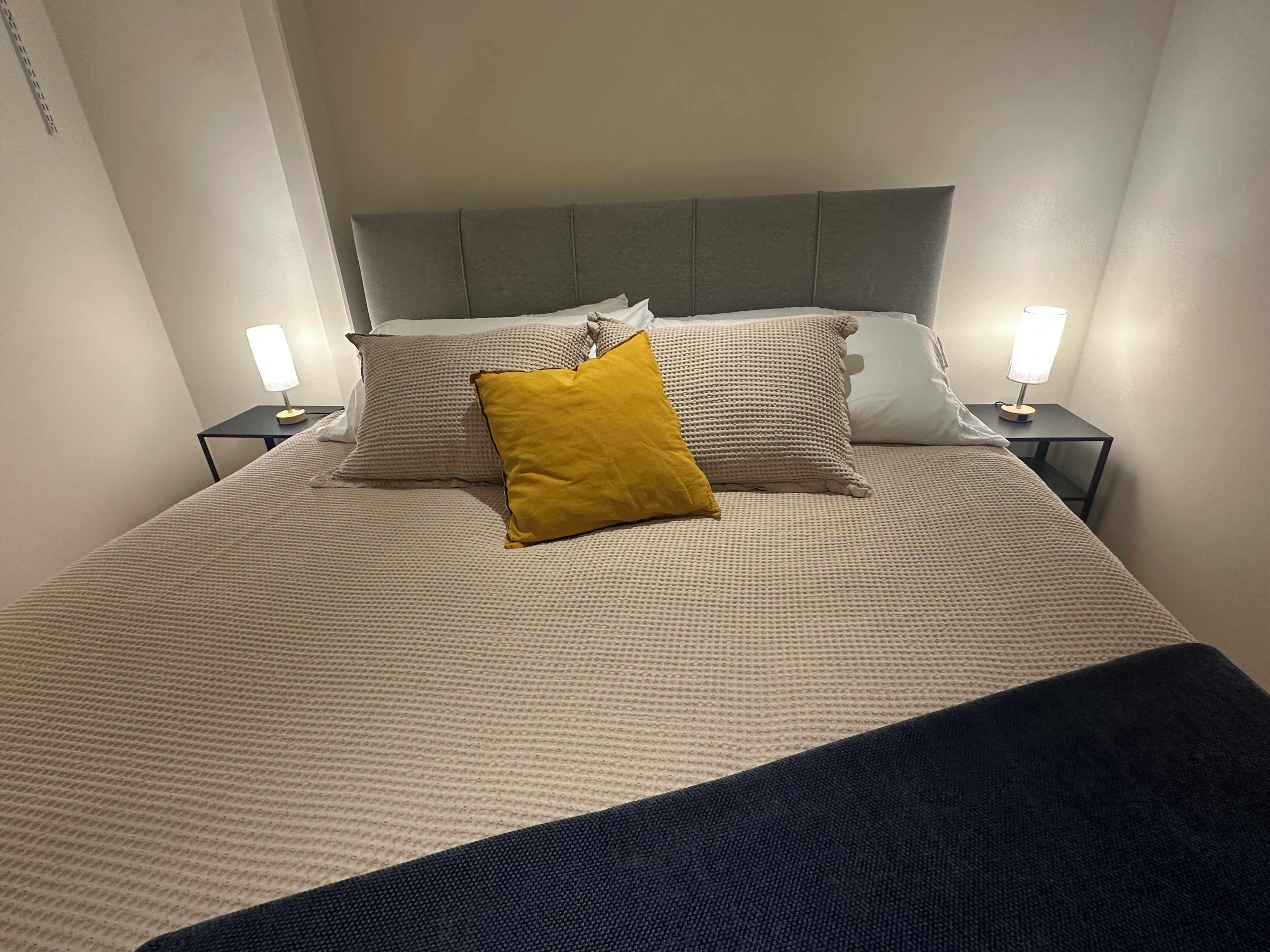 Ein ordentlich bezogenes Bett mit grauem Kopfteil, zwei Nachttischlampen und einem gelben Kissen.