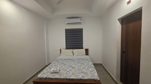 Una habitación estrecha con una cama, un ventanal con persianas, un equipo de aire acondicionado y una puerta.
