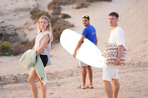 Three individuals on a sandy beach, one holding a surfboard and another carrying a beach bag.