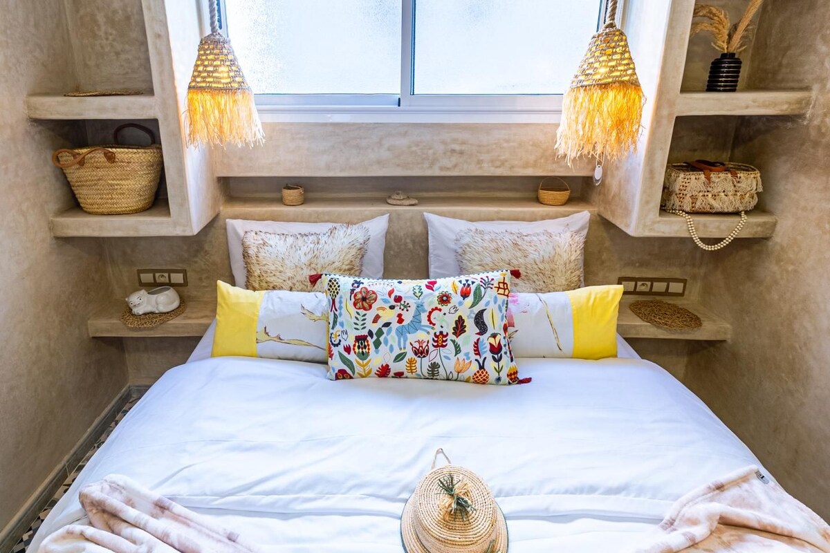 A bed with a colorful floral pillow, a woven basket, and a woven hat on the bed. There are two hanging lamps with tassels, and shelves with baskets and a small figurine.