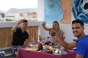 A group of people seated at a table with food and drinks, a map on the wall, and a view of the cityscape. - Waves Hunter Surf House - Anza (Agadir)