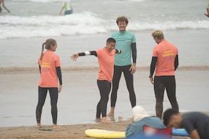 Un grupo de personas en una playa con tablas de surf, algunas vestidas con camisetas naranjas con el texto 'Waves Hunter'.