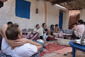 Un grupo de personas sentadas en el suelo en un entorno rústico al aire libre, con un techo de paja y rodeadas de mesas con diversos objetos.