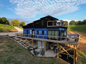 A two-story building with a blue exterior, a deck with outdoor seating, and a dirt road in the background. - Waterfall Cabin - Hot Tub-Cozy Theater & Game Room W/ Surround Sound & Nintendo Switch 2 - Griddle Grill - Outdoor TV area with Swings, Hammocks, & Fire pit! (Denniston)
