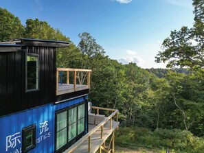 A two-story building with a balcony, surrounded by dense forest. - Waterfall Cabin - Hot Tub-Cozy Theater & Game Room W/ Surround Sound & Nintendo Switch 2 - Griddle Grill - Outdoor TV area with Swings, Hammocks, & Fire pit! (Denniston)