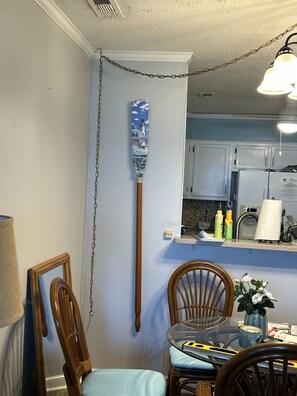 A dining area with a table and chairs, a kitchen in the background, and a decorative item on the wall. - Chateau By The Sea-Oceanfront-Sleeps4-2b/2b-Laundry-Kitchen-Pool-Free Parking (North Myrtle Beach)