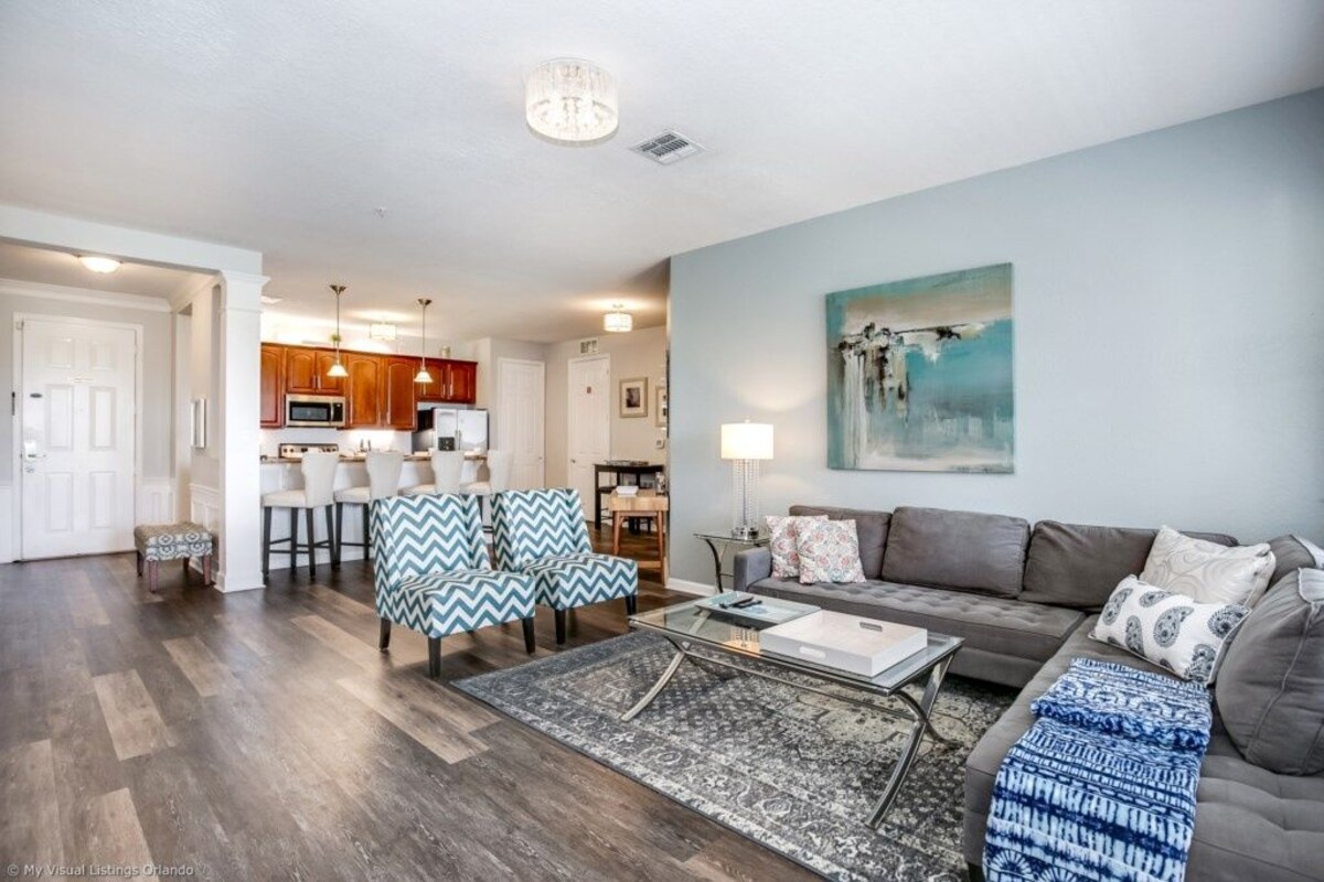Ein modernes Wohnzimmer mit grauem Sofa, gestreiftem Hocker und einem Couchtisch. Im Hintergrund befindet sich ein Essbereich mit Holzkohle und eine Küche.