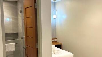 A modern hotel room with a glass shower enclosure, a wooden door, a bedside table with a lamp, and a bed.