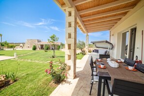 Ein überdachter Außenbereich mit Grill, Tisch und Stühlen und Blick auf ein Gebäude im Hintergrund.