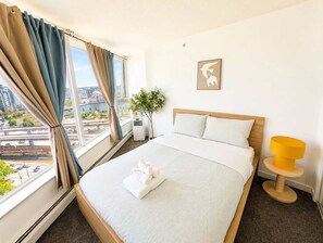 A hotel room with a bed, a window with curtains, a bedside table with a lamp, and a wall-mounted picture. - Luxe Retreat | BCPlace/Skytrain/View/Pool/Jacuzzi (Vancouver)