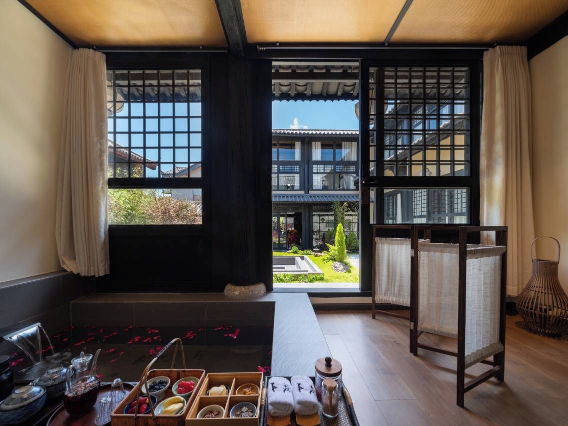 A room with tatami flooring, a low table with a tea set, a view of a traditional building through a shoji screen, and a woven basket.