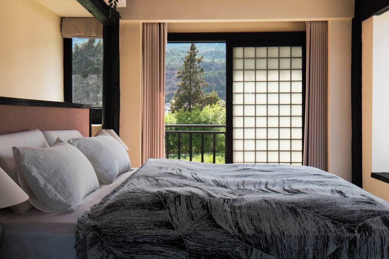 A hotel room with a bed, a view of a balcony, and a window with a view of trees and mountains.