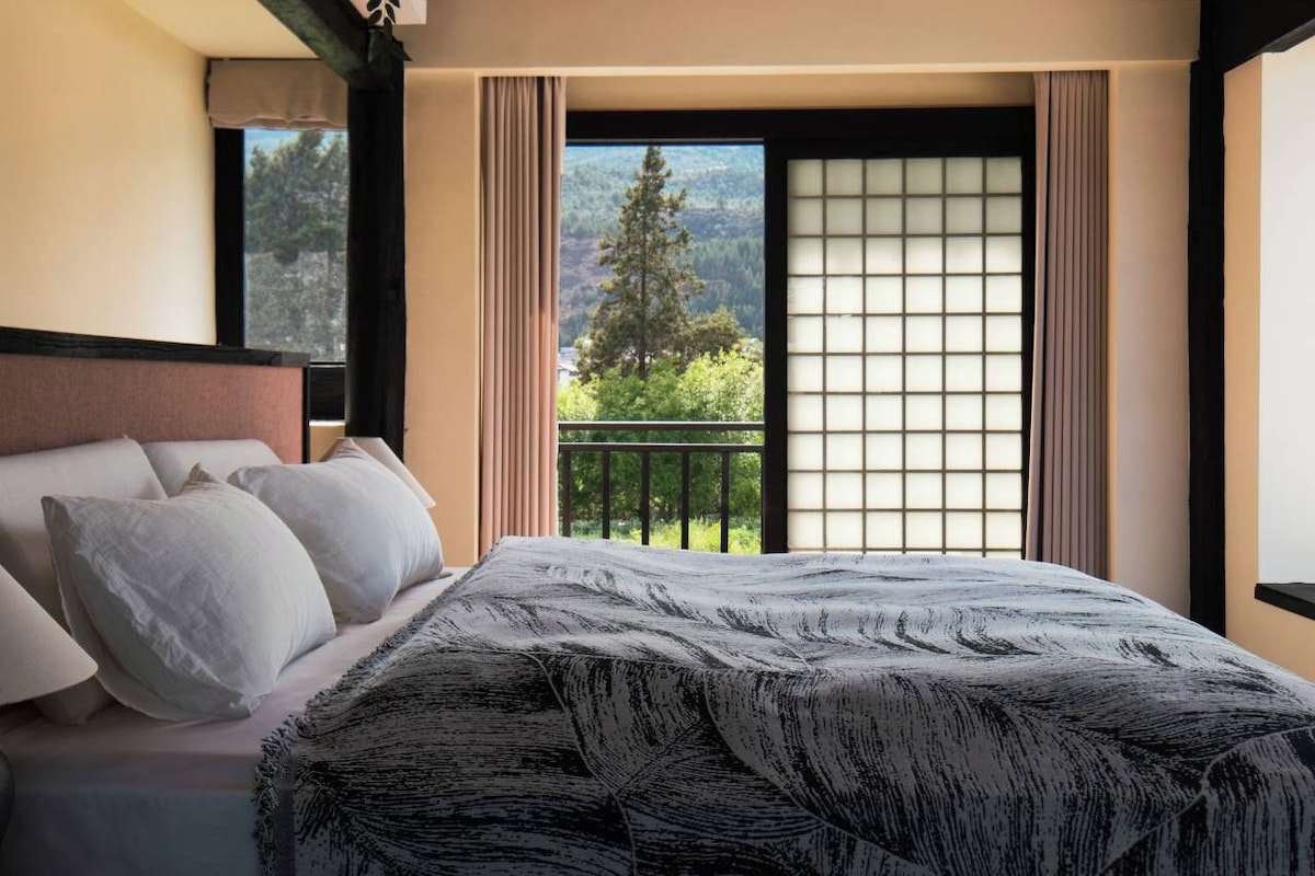 A hotel room with a bed, a view of a balcony, and a window with a view of trees and mountains.