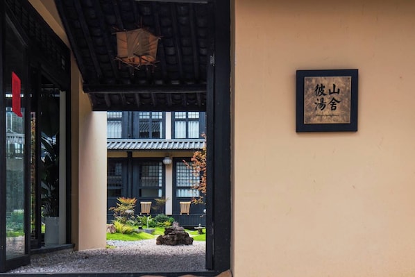 A traditional Japanese building entrance with a black gate, a framed sign with Japanese characters, and a view of a courtyard with a rock, plants, and a tiled roof.