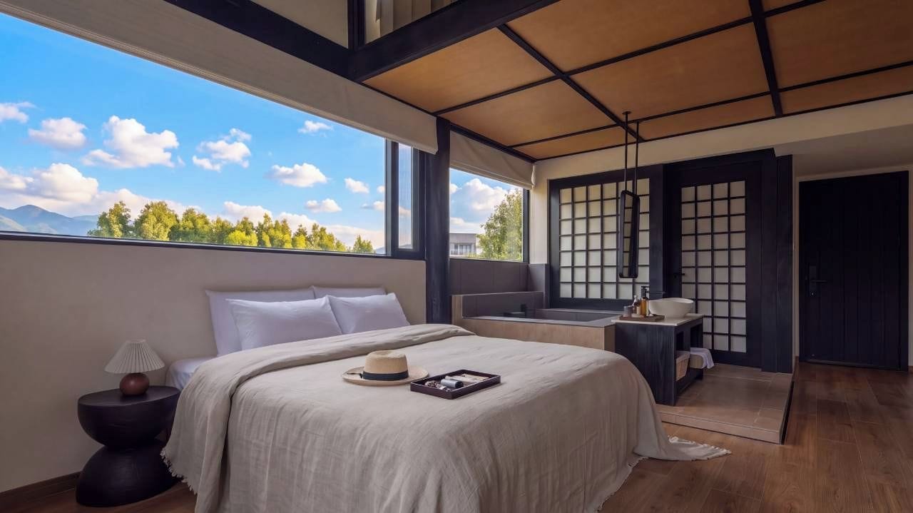 A modern bedroom with a large bed, a small table, a window with a view of trees and mountains, and a sliding door.