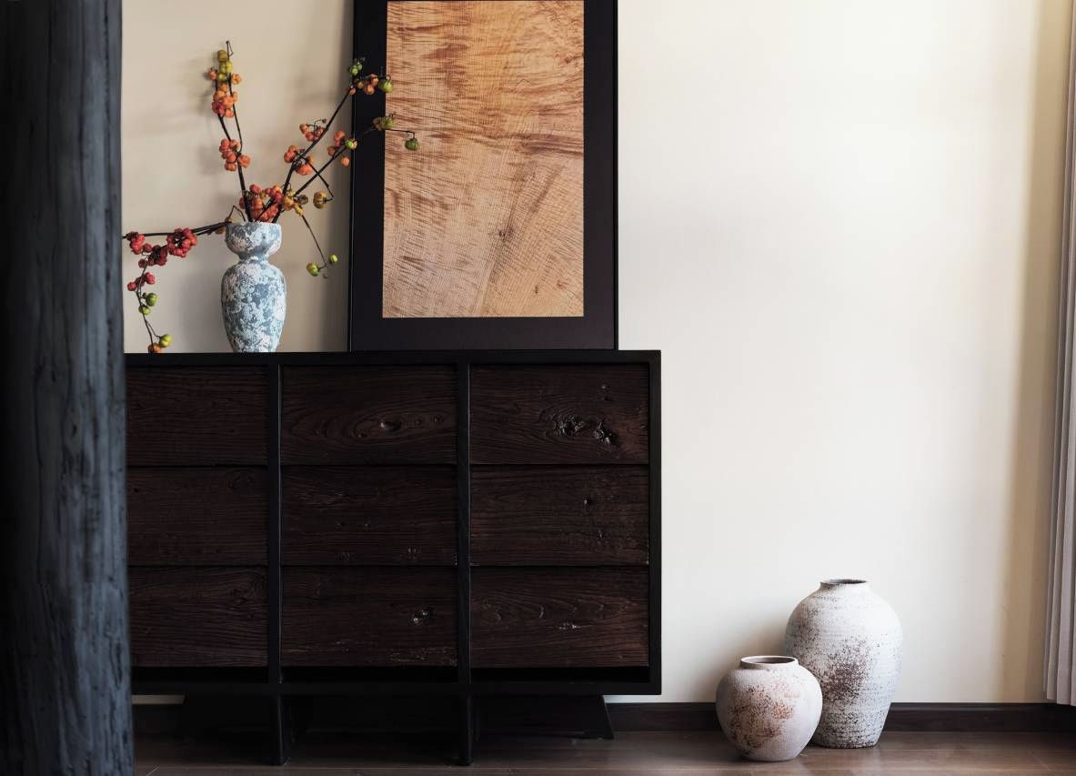 Un mueble de madera oscura con una obra de arte enmarcada encima, dos jarrones de cerámica en el suelo junto al mueble, y un jarrón con ramas sobre el mueble.