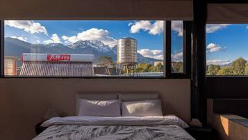 Un dormitorio con cama, almohadas, mesita de noche y ventana con vista a montañas y una torre de agua.