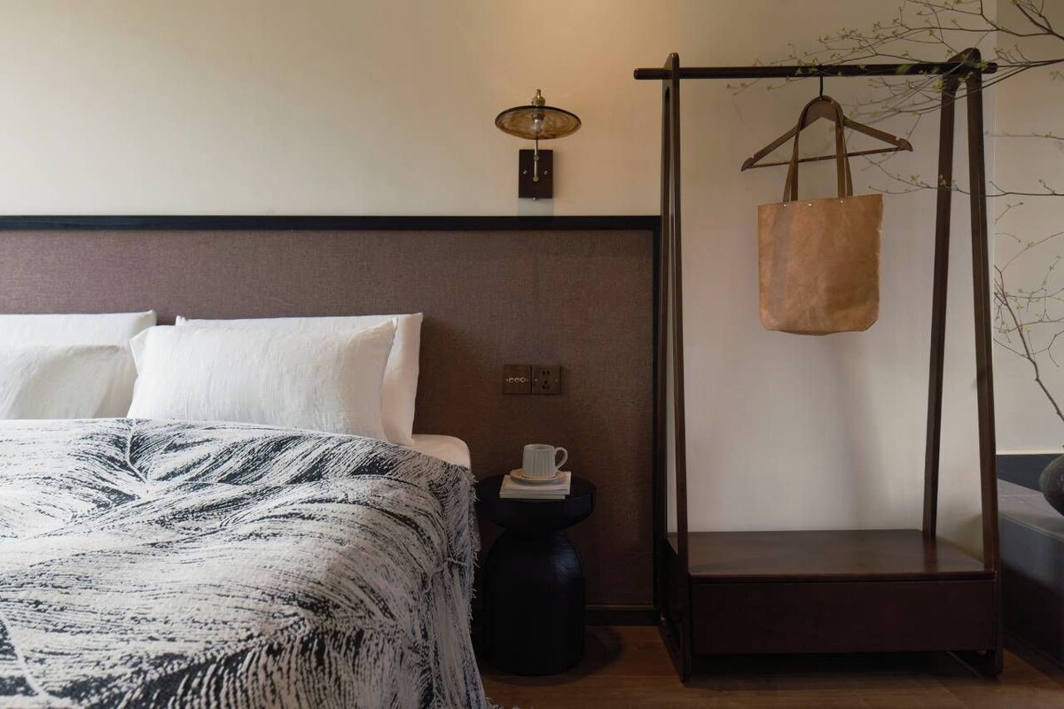 A bed with a patterned bedspread, a bedside table with a cup and book, a hanging bag, and a wall-mounted lamp.
