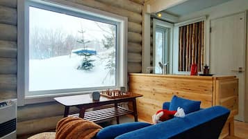 A cozy cabin interior with a large window, a wooden table with chess pieces, a blue chair, and a small dog.