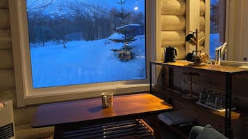A cozy cabin interior with a large window overlooking a snowy landscape, a wooden table, and a kitchen area with a kettle and mugs.