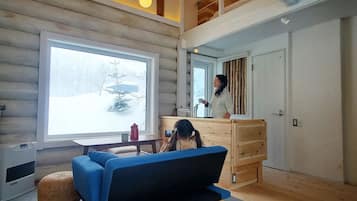 A cozy cabin interior with a sofa, a wooden dresser, and a large window with a snowy view.