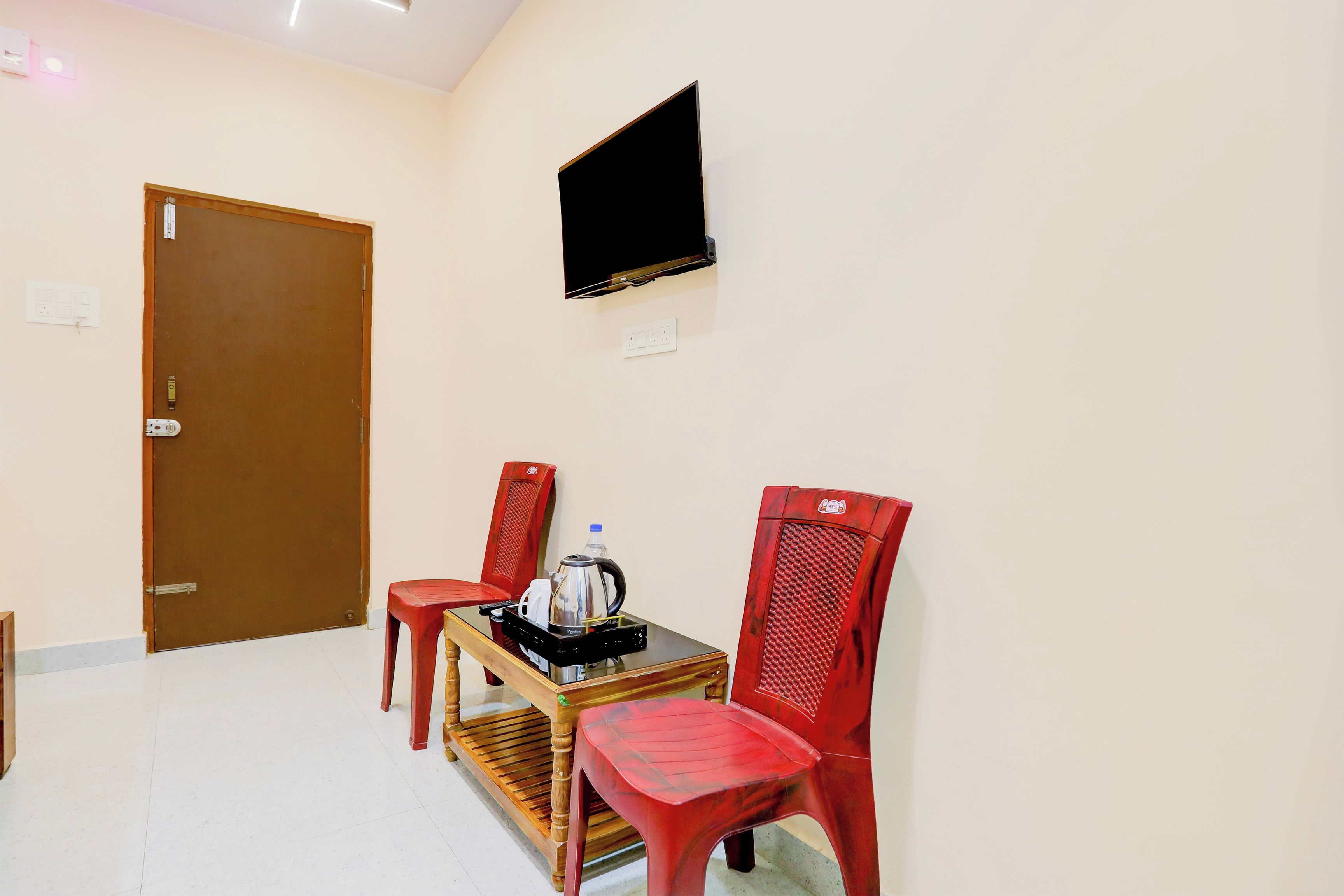 Una habitación con puerta de madera, televisor montado en la pared y dos sillas de plástico rojas alrededor de una pequeña mesa de madera con una tetera y una botella.