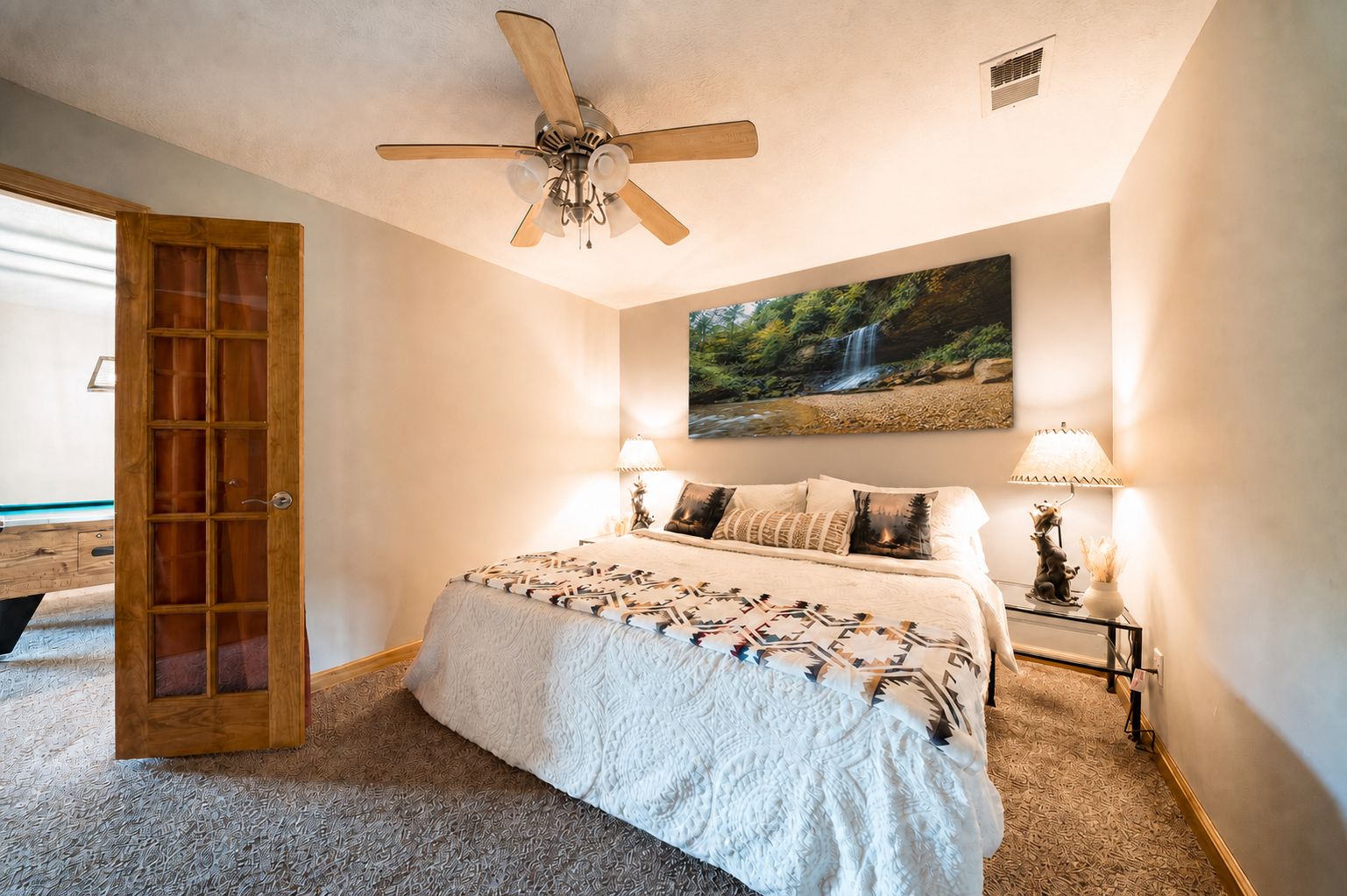 A bedroom with a bed, a ceiling fan, a painting of a waterfall, two bedside lamps, and a wooden door.