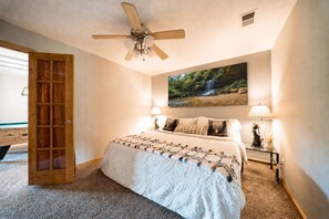 A bedroom with a bed, a ceiling fan, a painting of a waterfall, two bedside lamps, and a wooden door.