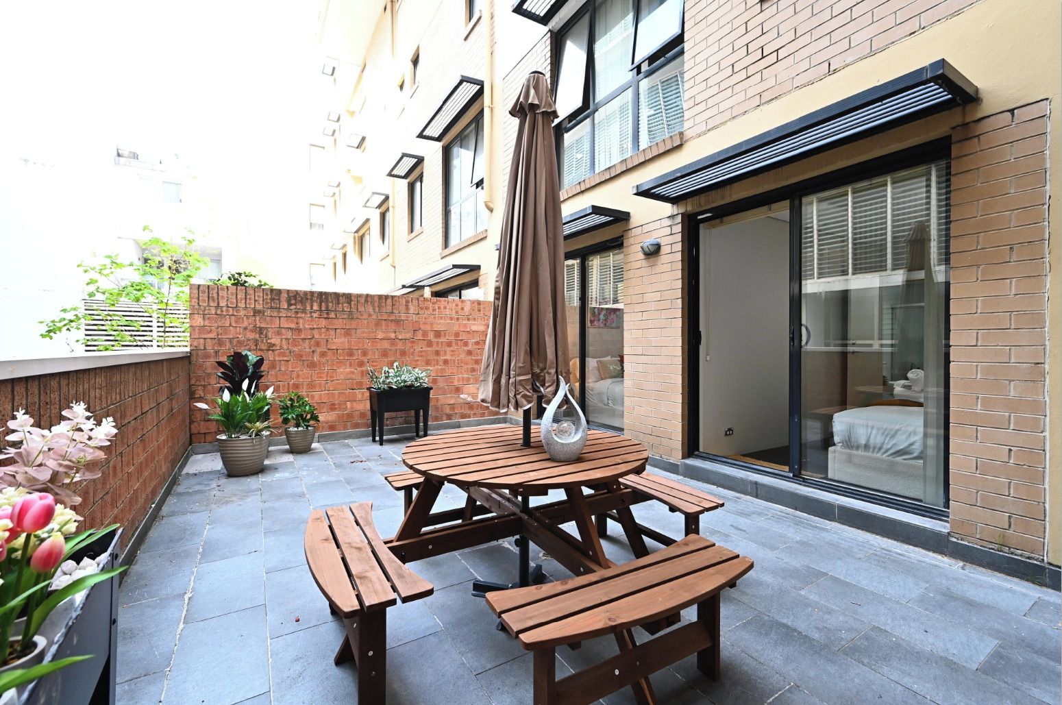 A patio with a wooden table and benches, potted plants, and a sliding glass door leading to a bedroom.