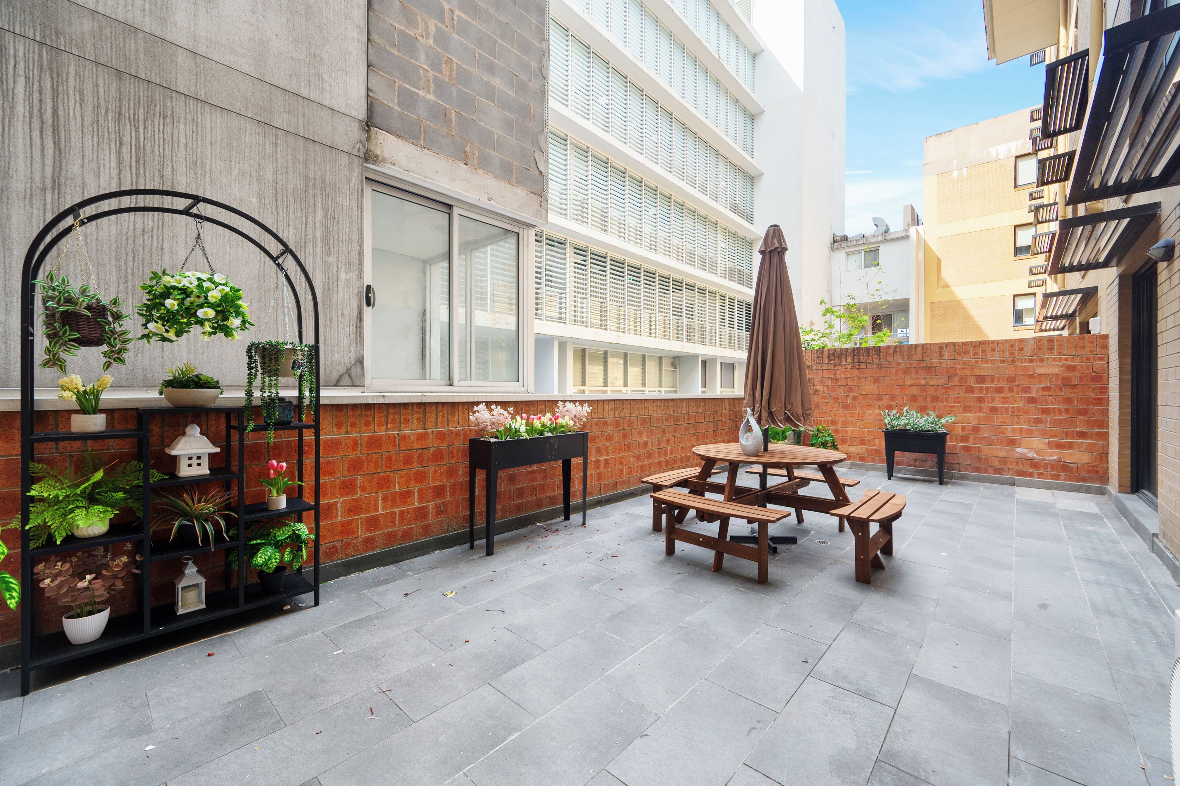 Un espace patio avec une table et des bancs en bois, entouré de plantes en pot et d’un mur de briques.