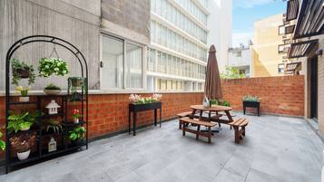 Un espace patio avec une table et des bancs en bois, entouré de plantes en pot et d’un mur de briques.