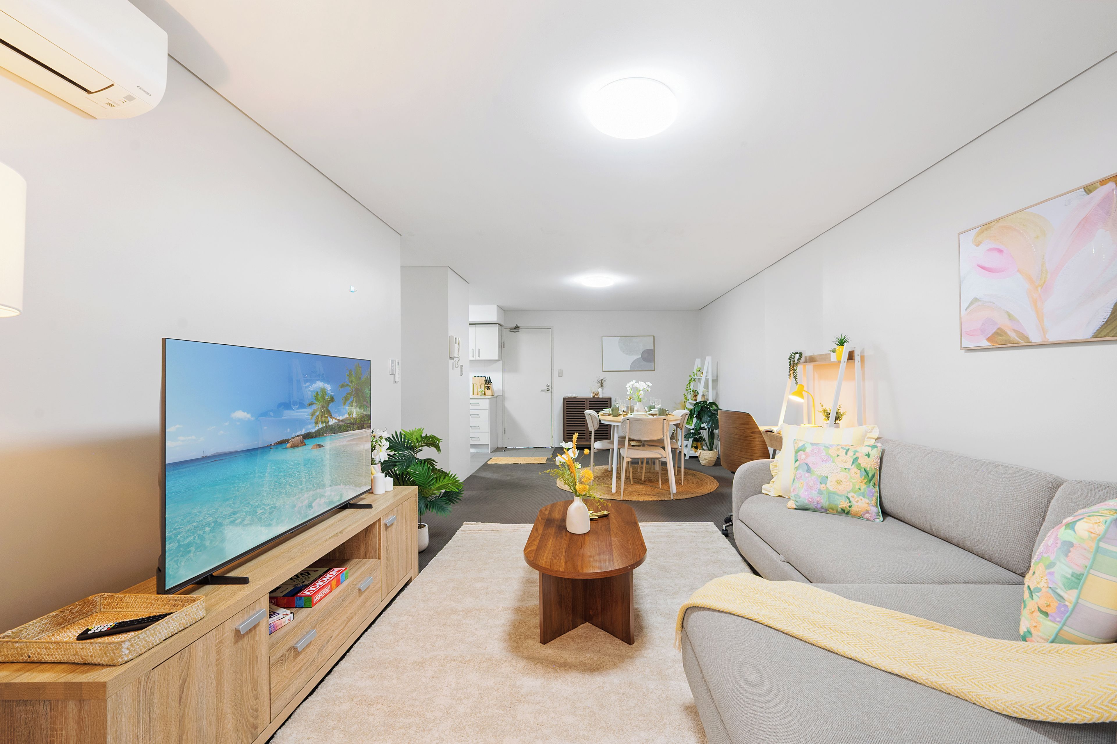A modern living room with a flat-screen TV, a wooden coffee table, a grey sofa, and a dining area in the background.