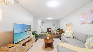 A modern living room with a flat-screen TV, a wooden coffee table, a grey sofa, and a dining area in the background.