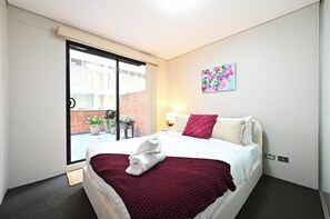 Habitación de hotel con cama, ventana, puerta, una obra de arte colgada en la pared y una mesita de noche con lámpara.