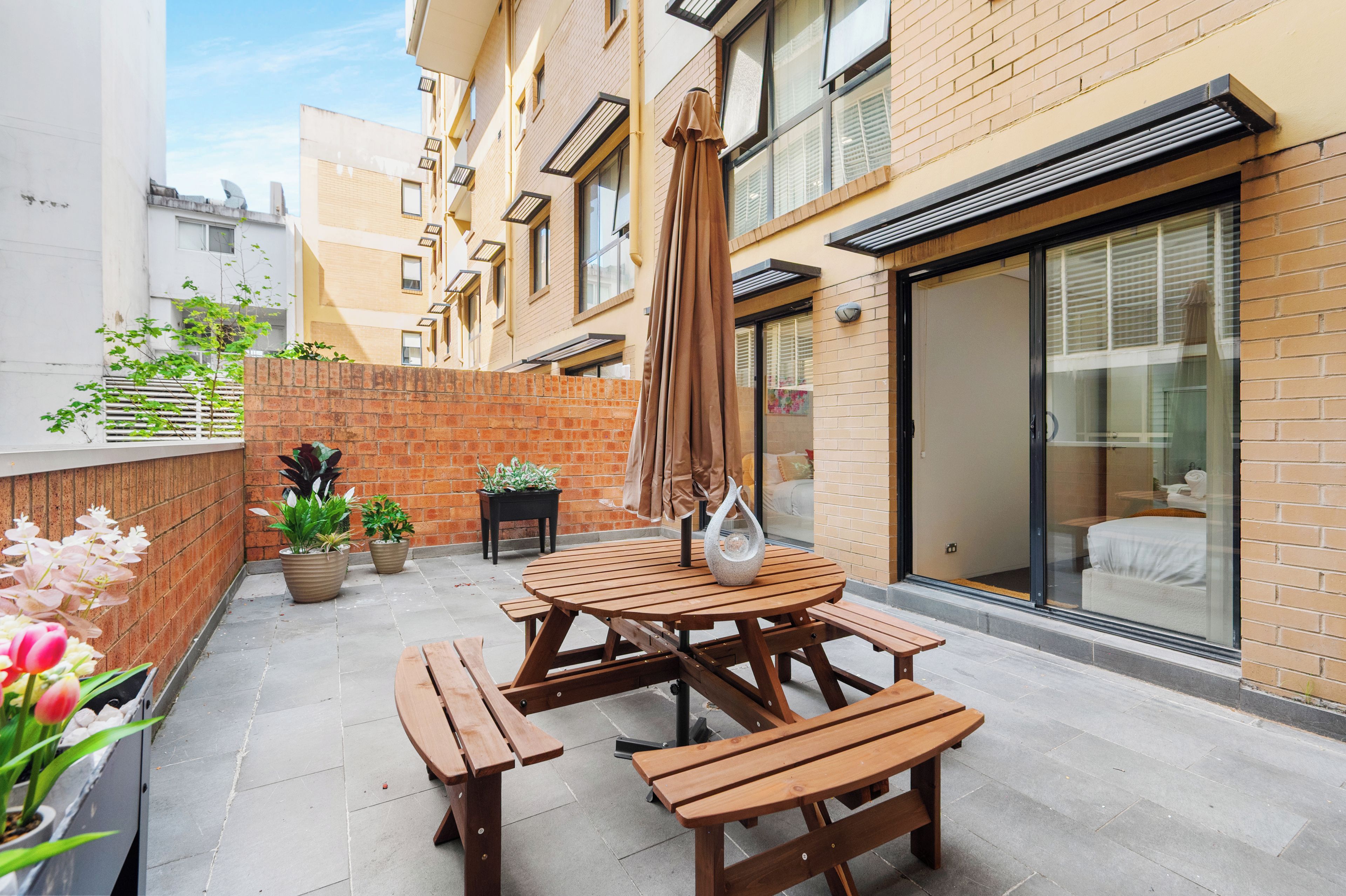 A patio with wooden tables and benches, potted plants, and a sliding glass door leading to a bedroom.