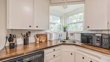 Une cuisine avec des meubles blancs, un plan de travail en bois, un lave-vaisselle, un évier et une fenêtre donnant sur des arbres.