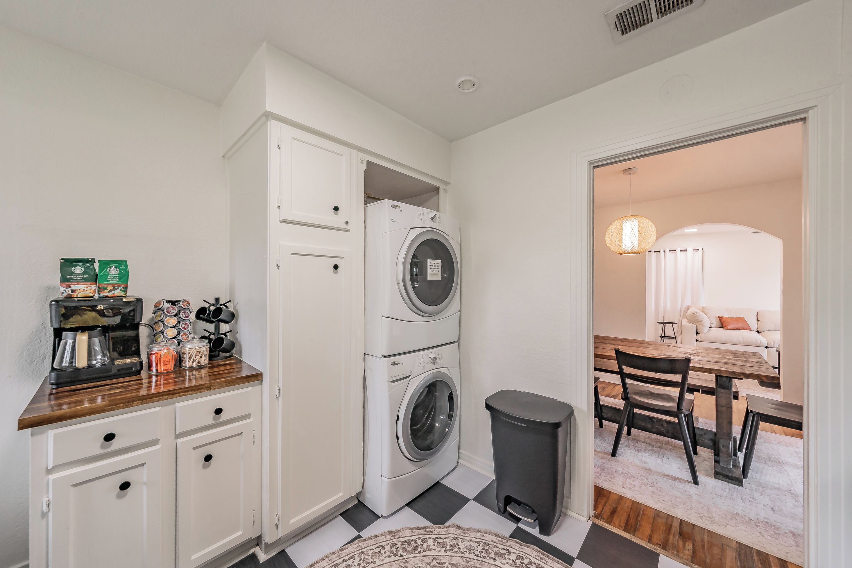 A compact kitchen with a coffee machine, storage cabinets, a washing machine, and a trash bin.