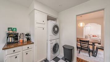 A compact kitchen with a coffee machine, storage cabinets, a washing machine, and a trash bin.