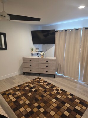 Ein Zimmer mit einem an der Wand befestigten Flachbildfernseher, einer Holzkommode mit Schubladen, einem gemusterten Teppich und einem Deckenventilator.