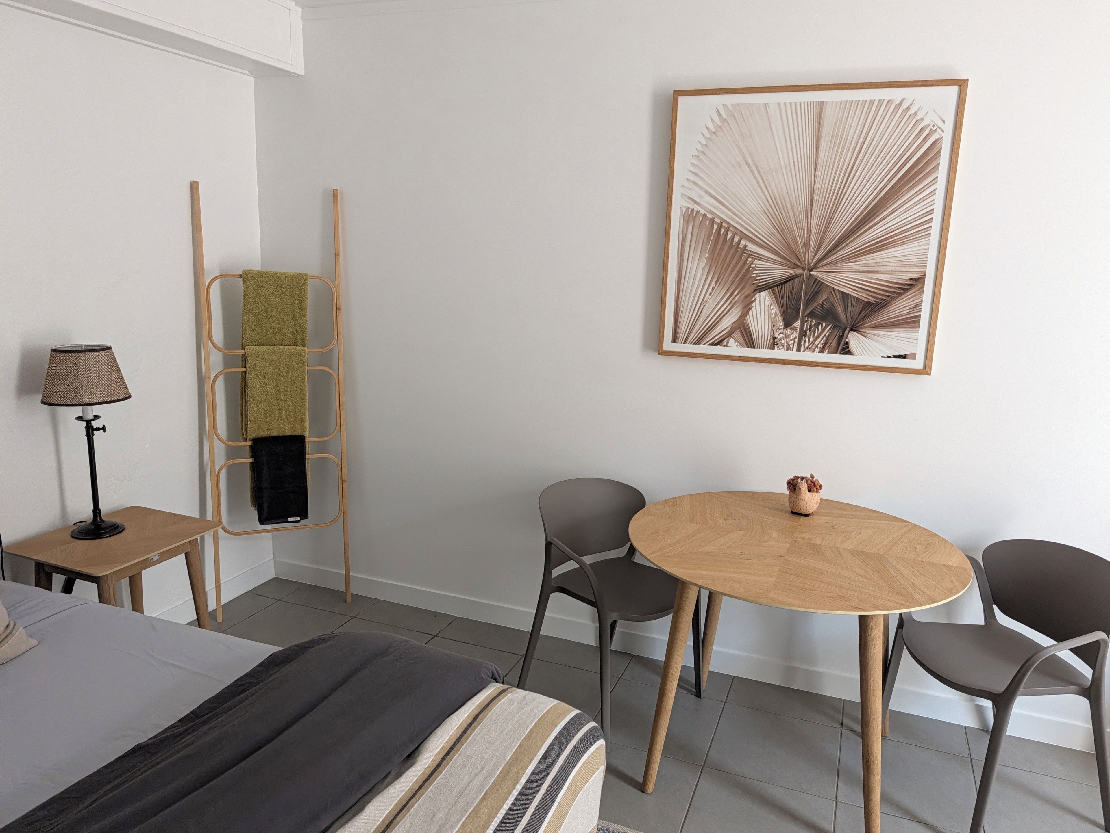 Et hotelværelse med en seng, et lille bord med en lampe, en stol, et rundt træbord og et billede i ramme på væggen.