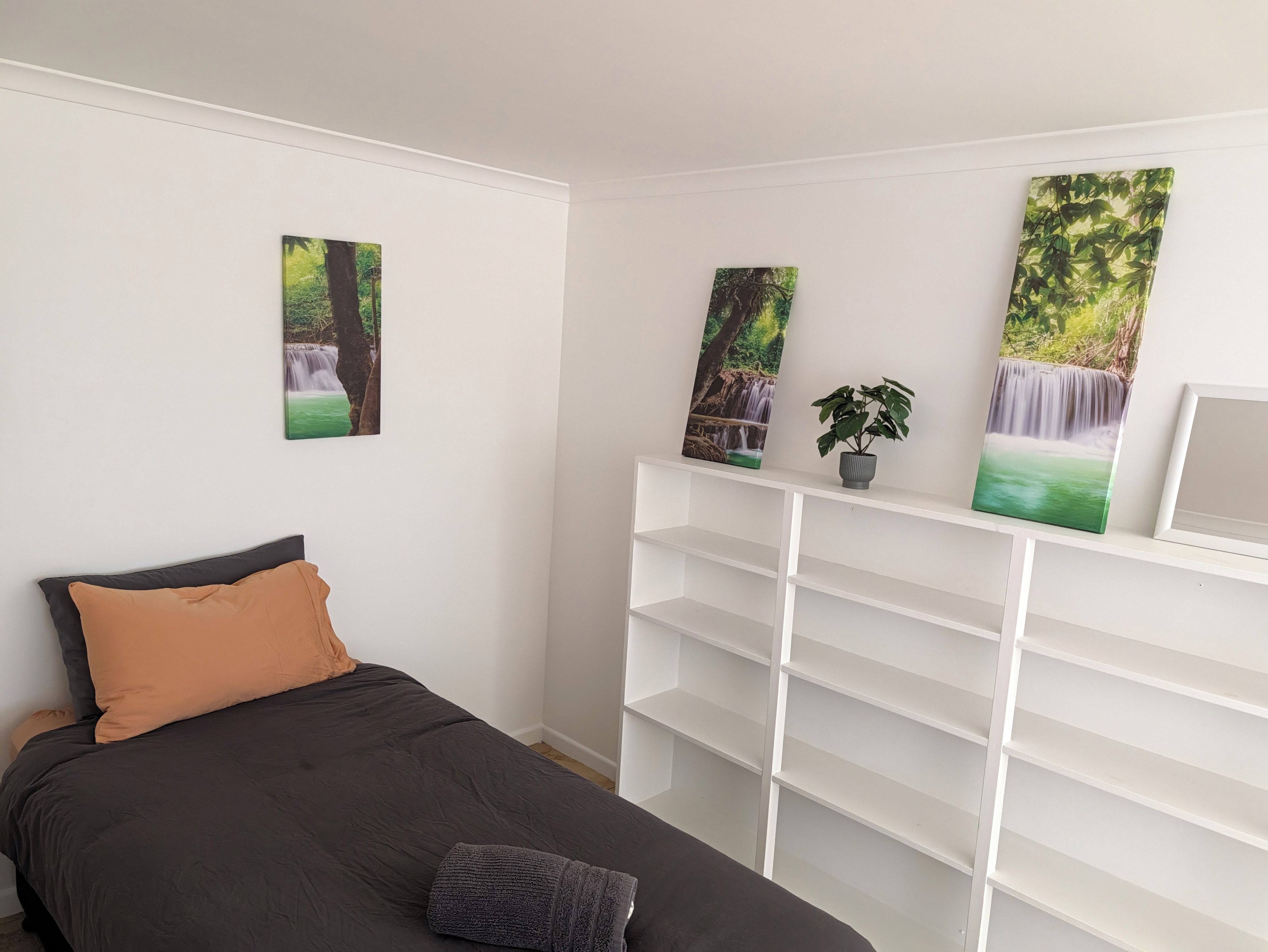 A bedroom with a bed, a white shelving unit, and three wall-mounted paintings.