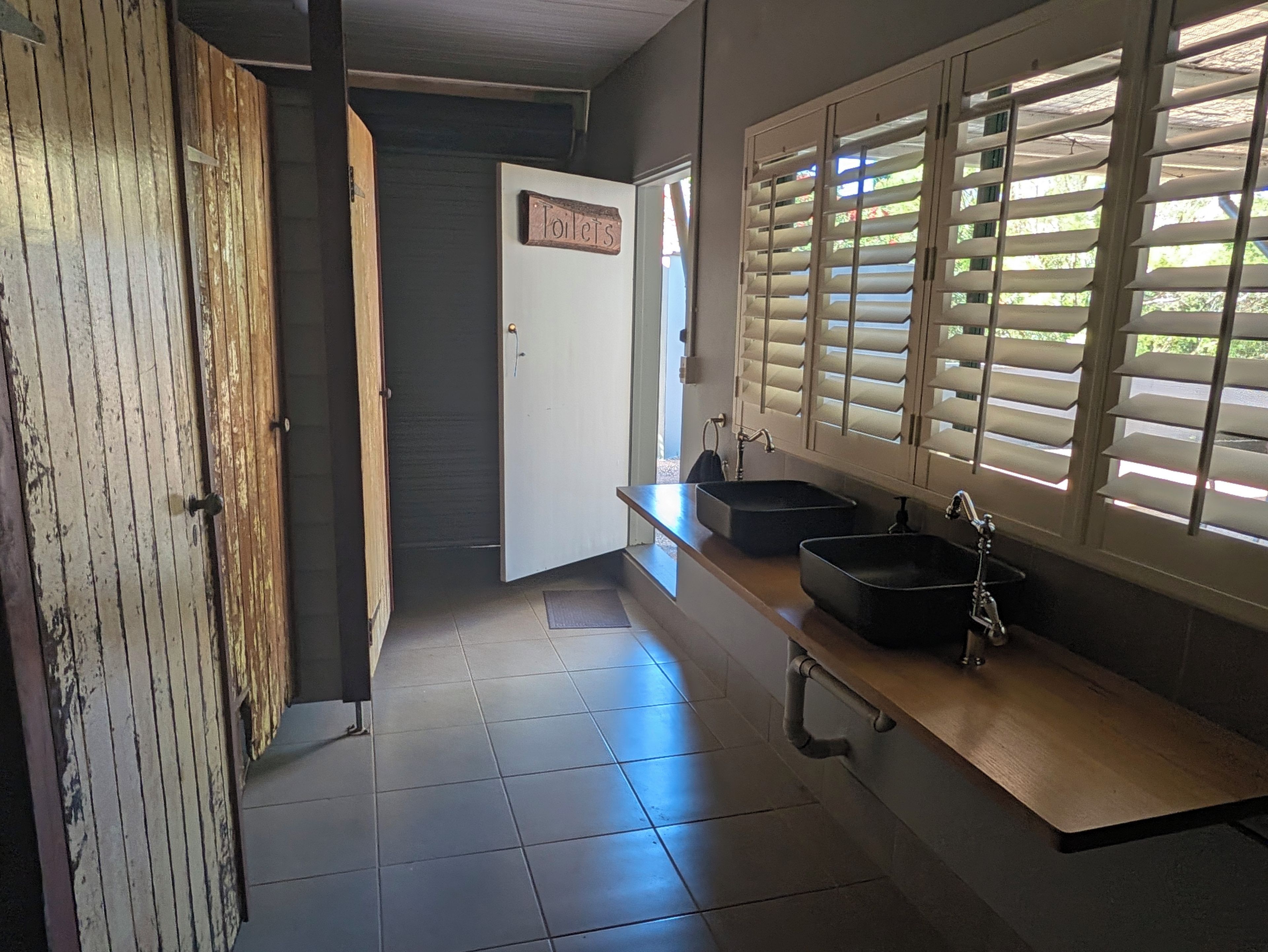 Ein Badezimmer mit Holztür, einem Schild mit der Aufschrift Toilette, einem Waschbecken mit Wasserhahn und einem Fenster mit Jalousien.