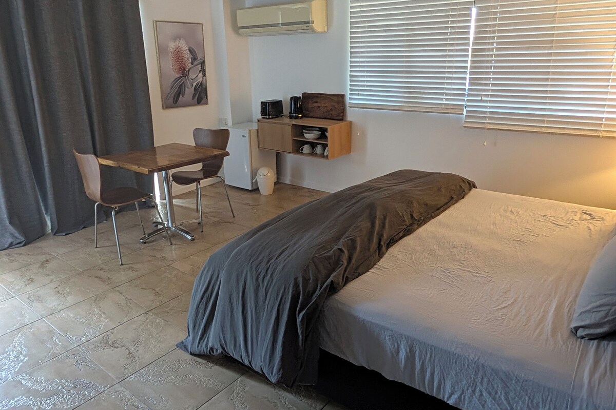A compact room with a bed, a desk, a chair, a small kitchenette, and a window with blinds.