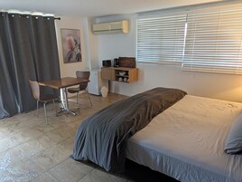 A compact room with a bed, a desk, a chair, a small kitchenette, and a window with blinds.